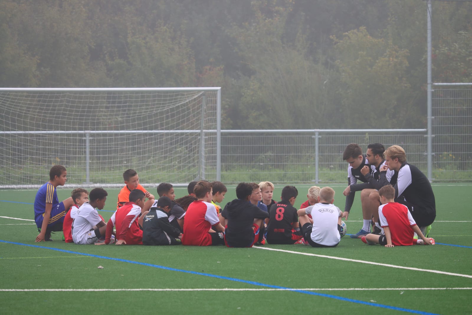 Spelers zitten in een kring op het veld terwijl de trainers uitleg geven