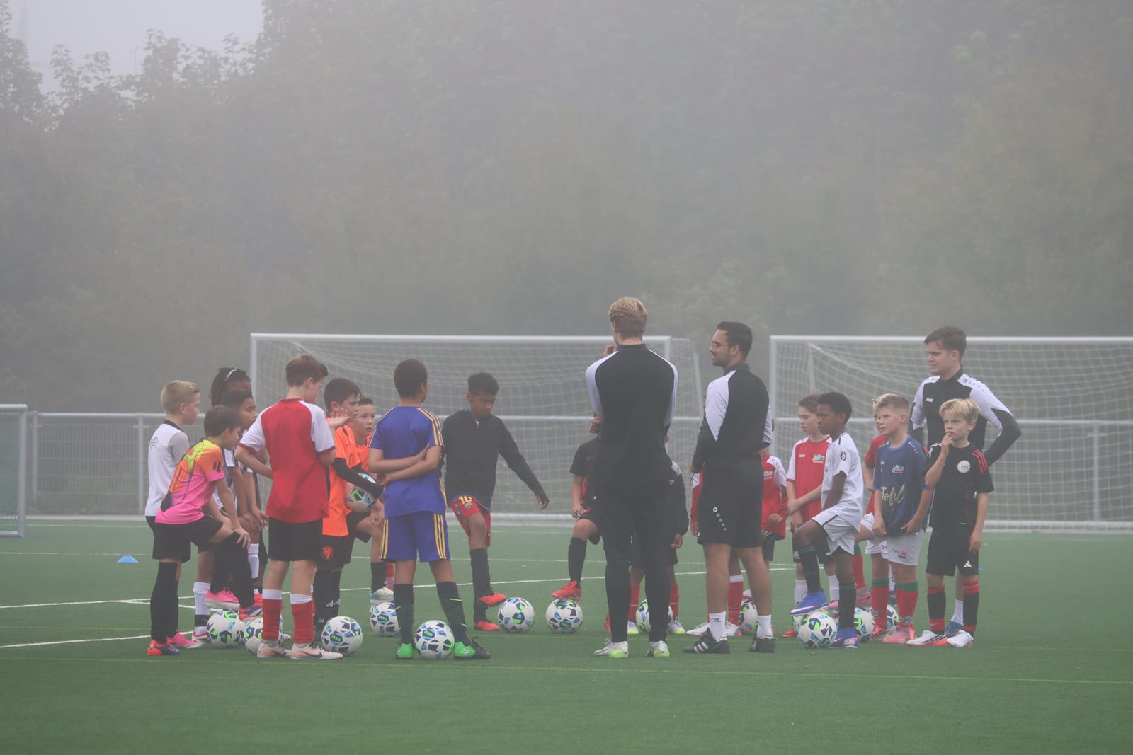 Spelers staan in de mist met elk een bal voor zich, de trainers geven instructies